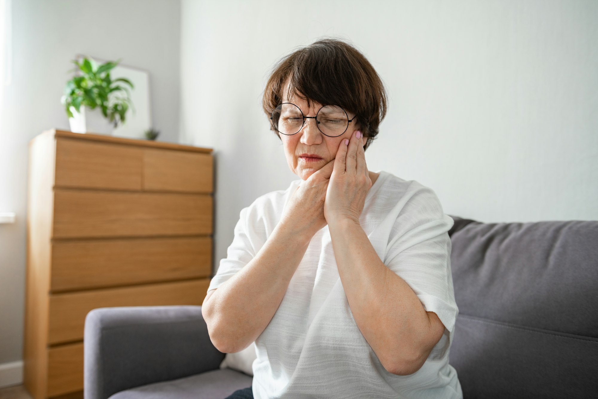 Elderly woman severe tooth pain sitting on the couch, health problems in old age